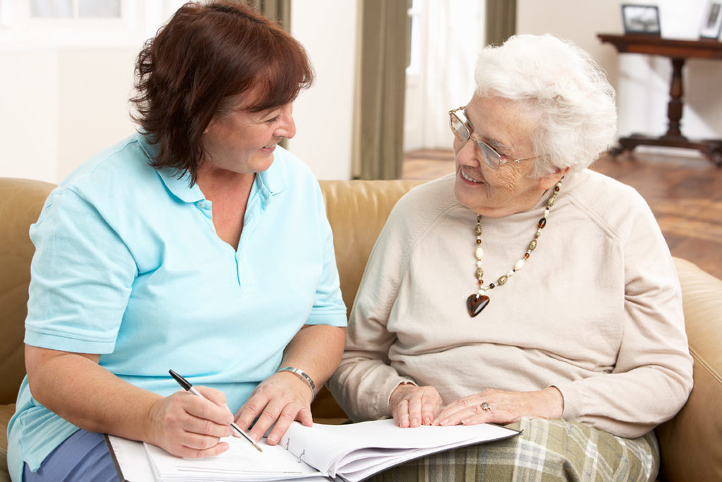 an image of a middle-aged woman helping her elderly mother fill out paper work as they both sit together on a couch inside a brightly lit room